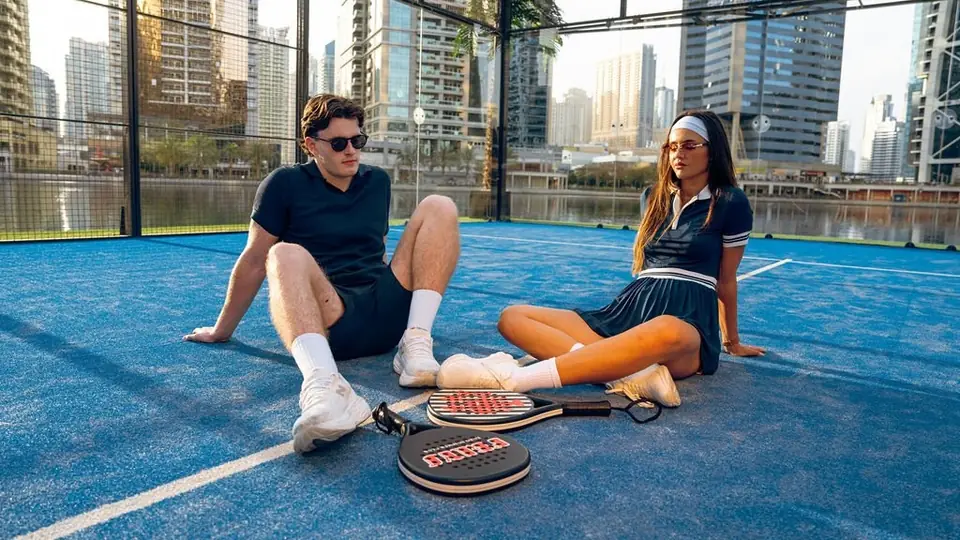 Two players in navy kit sitting on a blue padel court, their rackets on the ground between them.