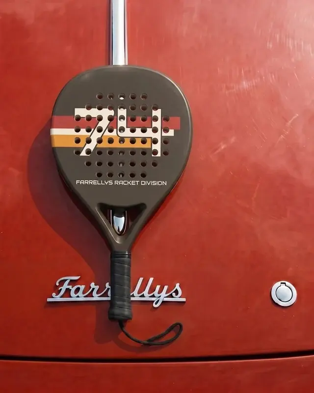 A charcoal padel racket with a striped 74 on its face, hung against red paintwork.