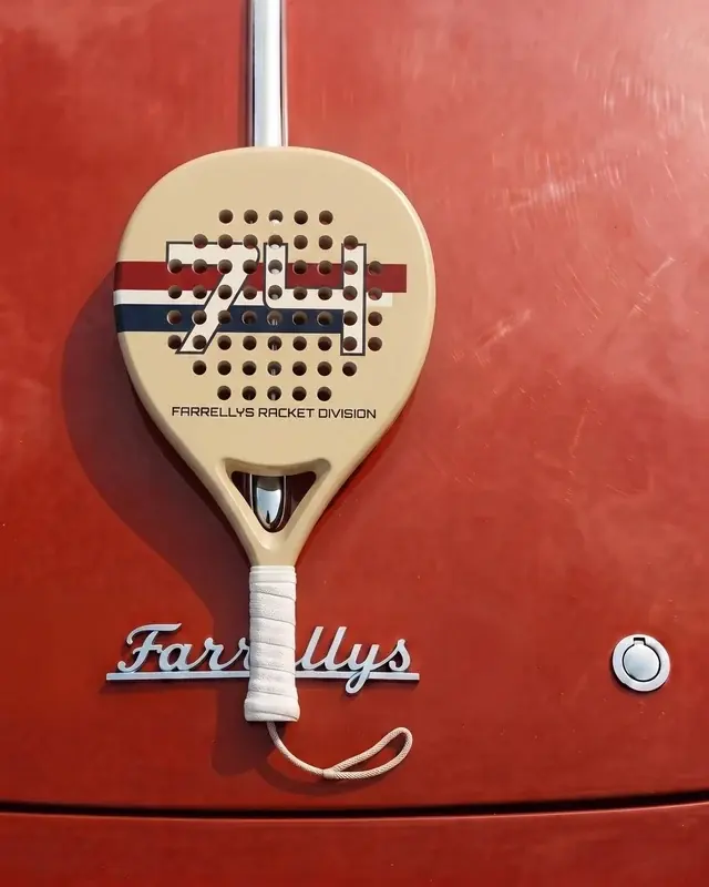A cream padel racket with a striped 74 on its face, hung against red paintwork.
