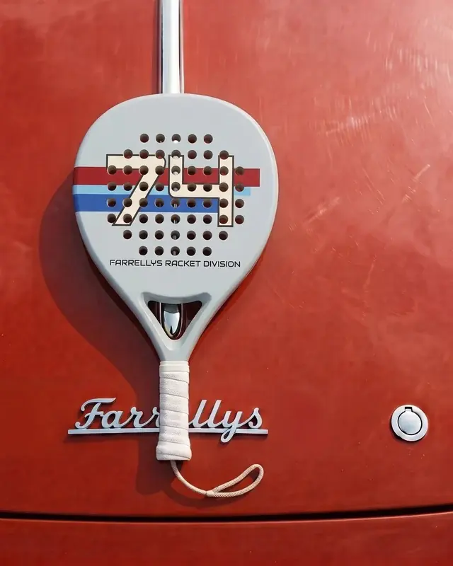 A light grey padel racket with a striped 74 on its face, hung against red paintwork.