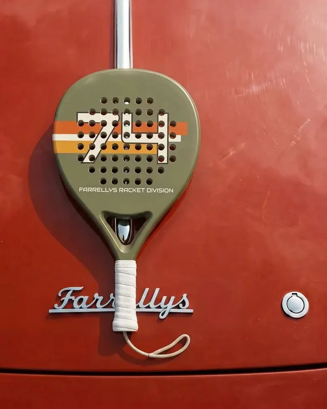 An olive padel racket with a striped 74 on its face, hung against red paintwork.